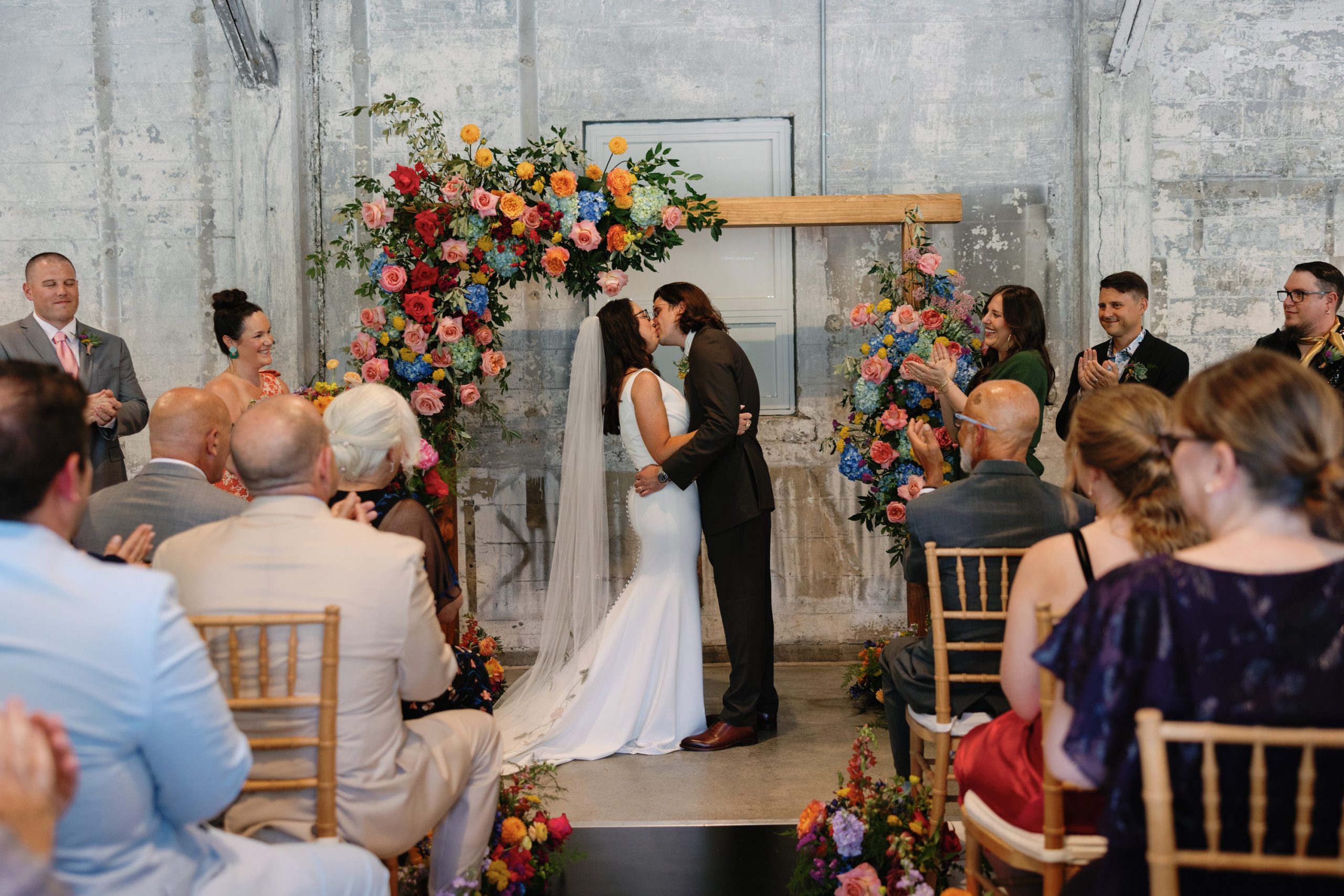 A bride and groom kiss under a wooden ceremony arch decorated with floral arrangements in front of seated friends and family members.