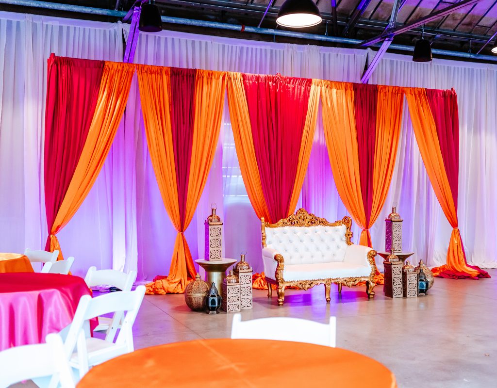 A curtained, pink-and-orange pipe-and-drape installation set against a white drape background, fronted by an ornate, gold-trimmed futon and golden-bronze lamps for a sangeet.