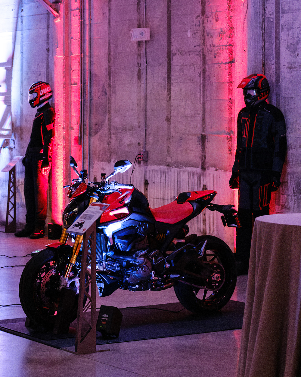 Two mannequins dressed in motorbike gear stand against a distressed stone garage wall with a Ducati bike in front of them.