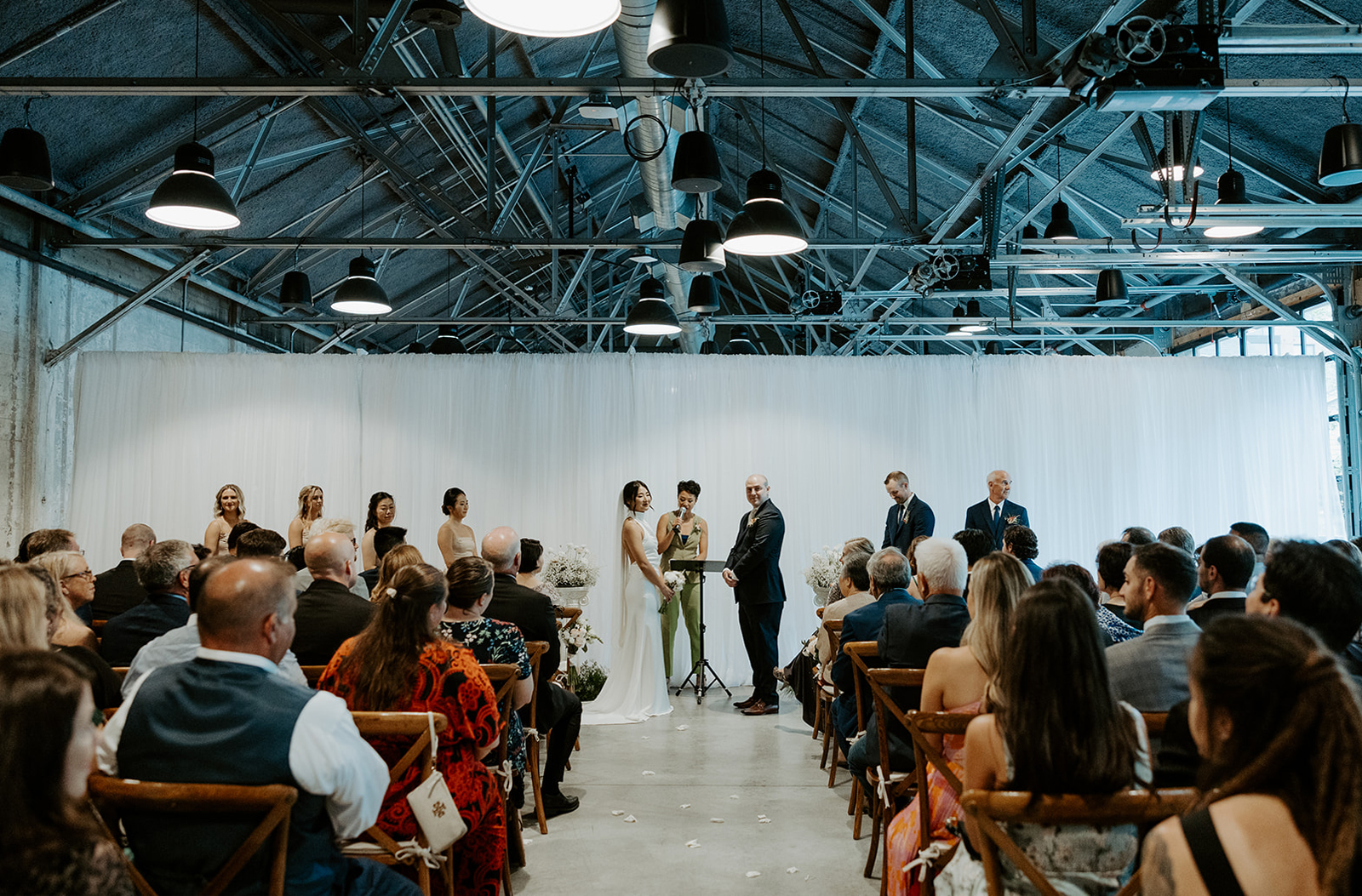 A bride and groom stand, holding hands, with an officiant in front of a wall-to-wall white pipe-and-drape installation with steel beams above them and their loved ones seated before them.