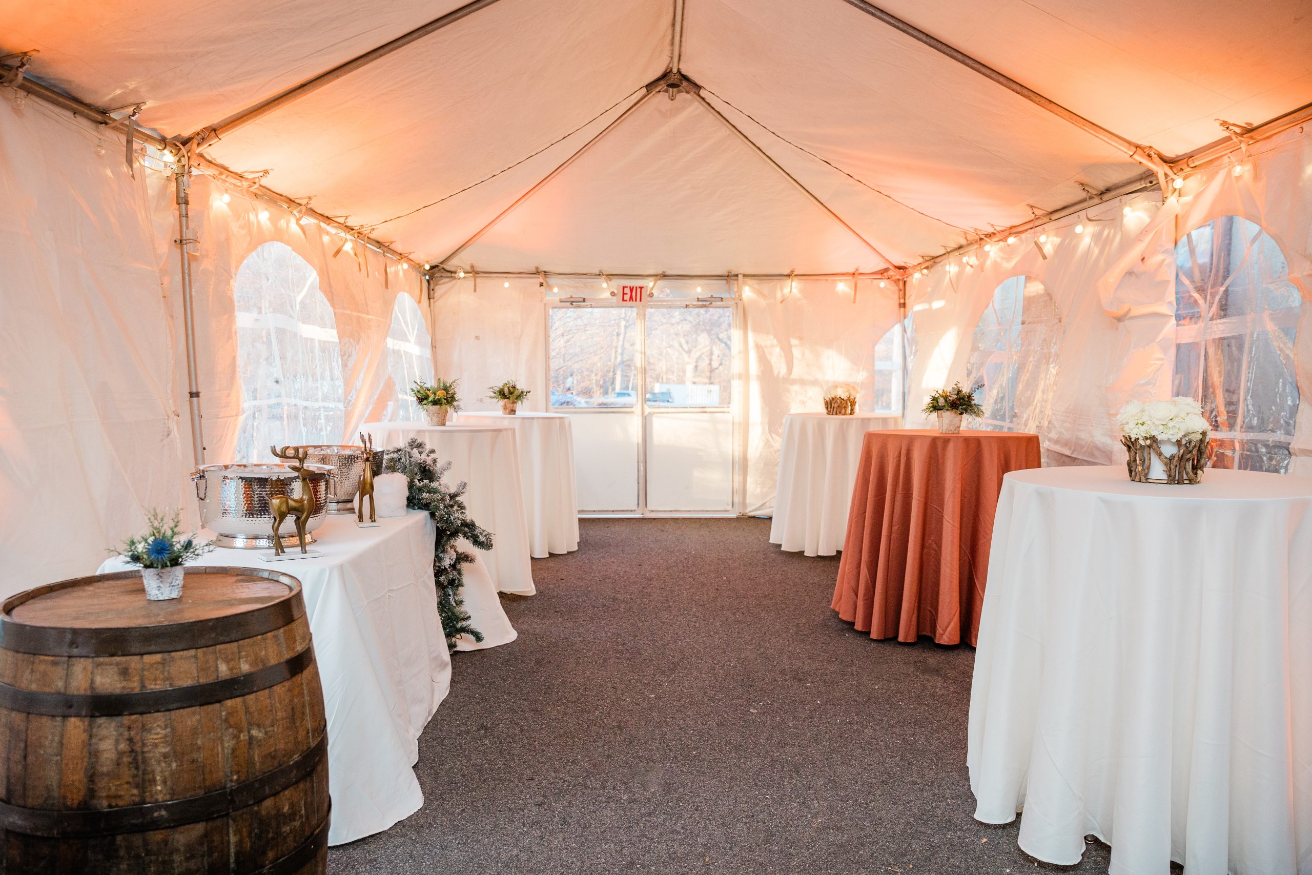 The interior of an outdoor event tent structure trimmed with warm string lights and outfitted with several high-top cocktail tables.