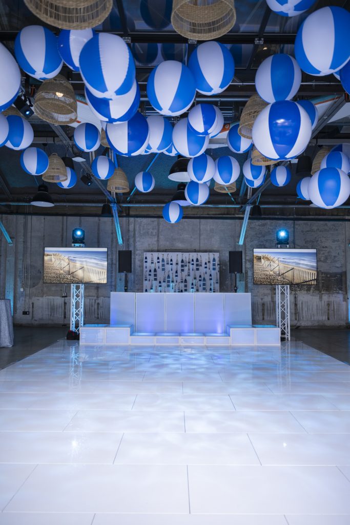 A DJ booth flanked by two monitors set against a distressed stone wall, fronted by a dance floor underneath a ceiling decorated with beach balls.