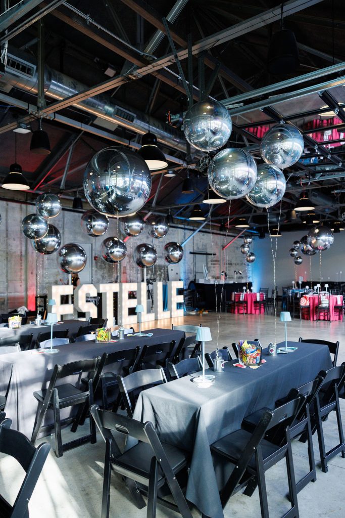 A bat mitzvah party setup in a warehouse-style venue, complete with a monogram in lights, reflective balloons, and black banquet tables and chairs.