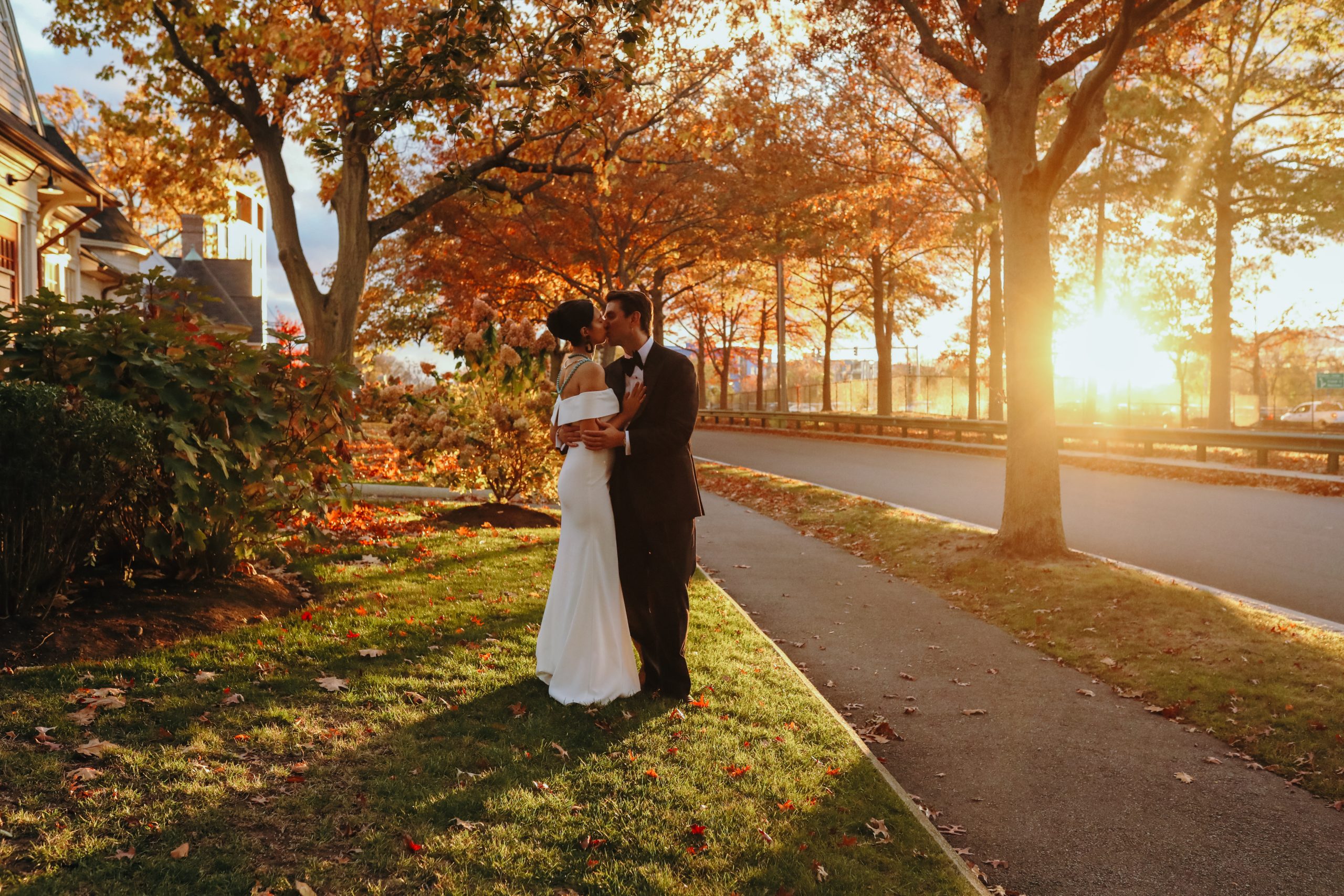 A bride and groom kiss on a roadside lawn as golden sunlight filters through fall foliage.