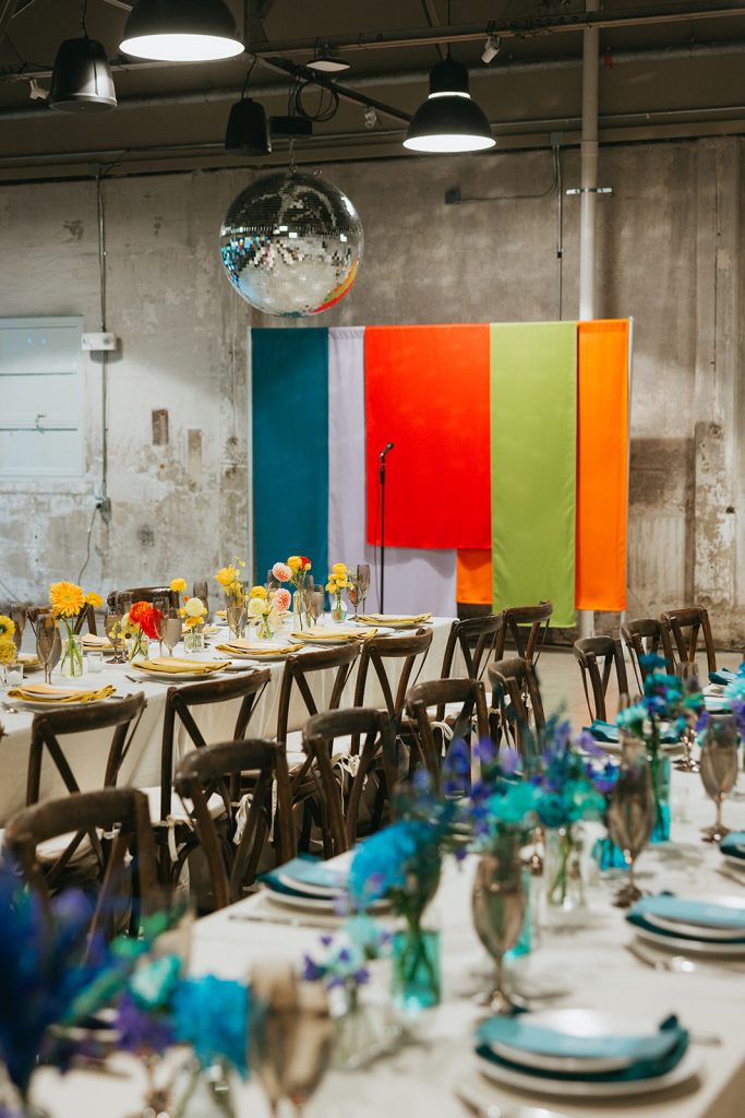 A backdrop of colorful sheets under a disco ball in front of long banquet tables lined with bud vases and rustic cross-back chairs set up for a wedding reception.