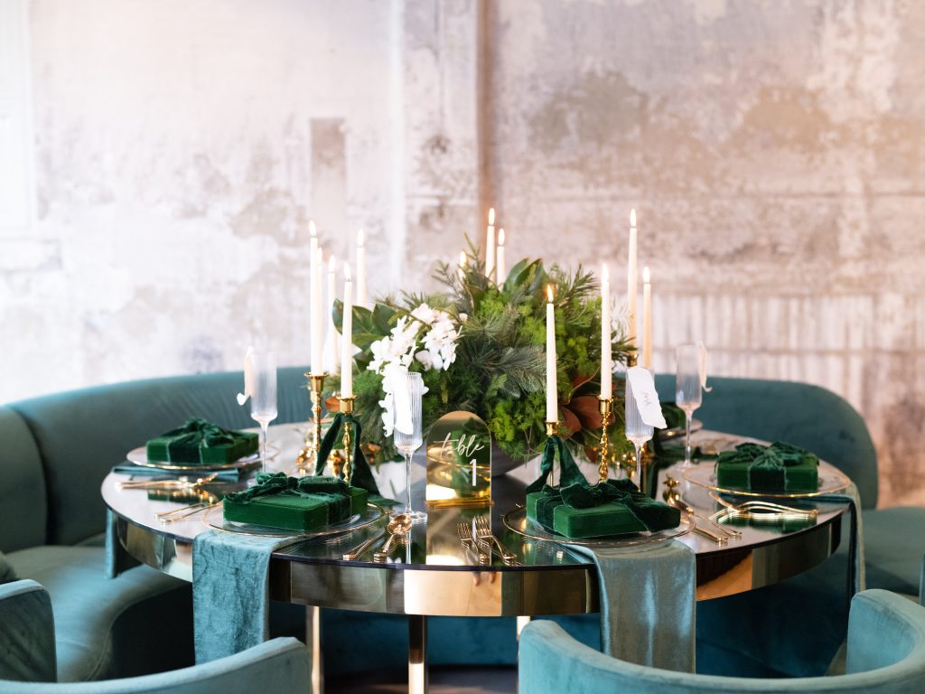 A formal round dining table design with a mixed fir centerpiece, taper candles, gilded dinnerware and flatware, and gift box decoration set against emerald velvet seating and a distressed stone wall.