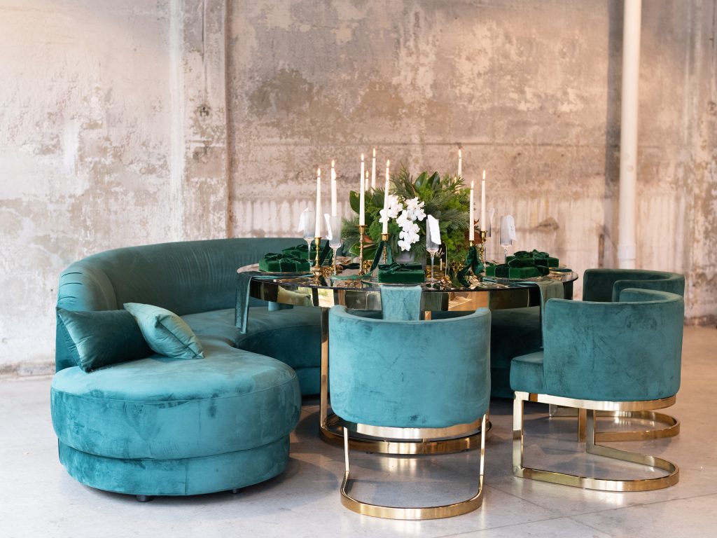 A formal round dining table design with a mixed fir centerpiece and taper candles surrounded by emerald velvet seating with gilded details set against a distressed stone wall.