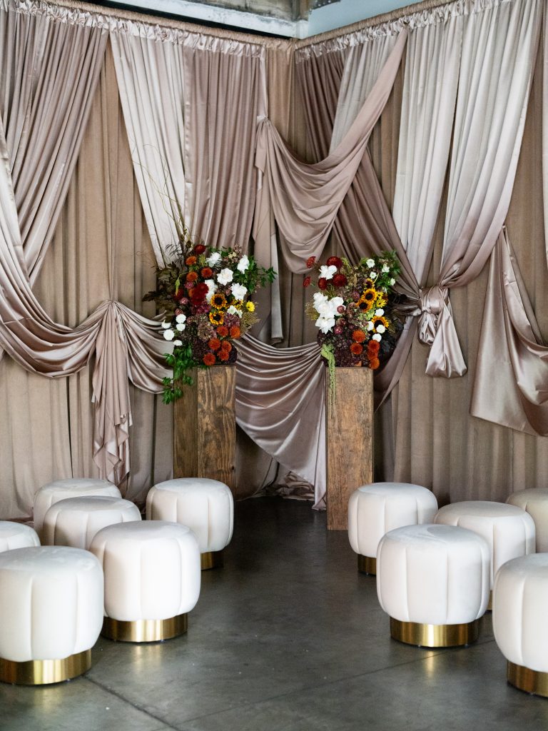 Two floral arrangements on wooden pedestals and a series of ottomans set against a designed drapery backdrop.