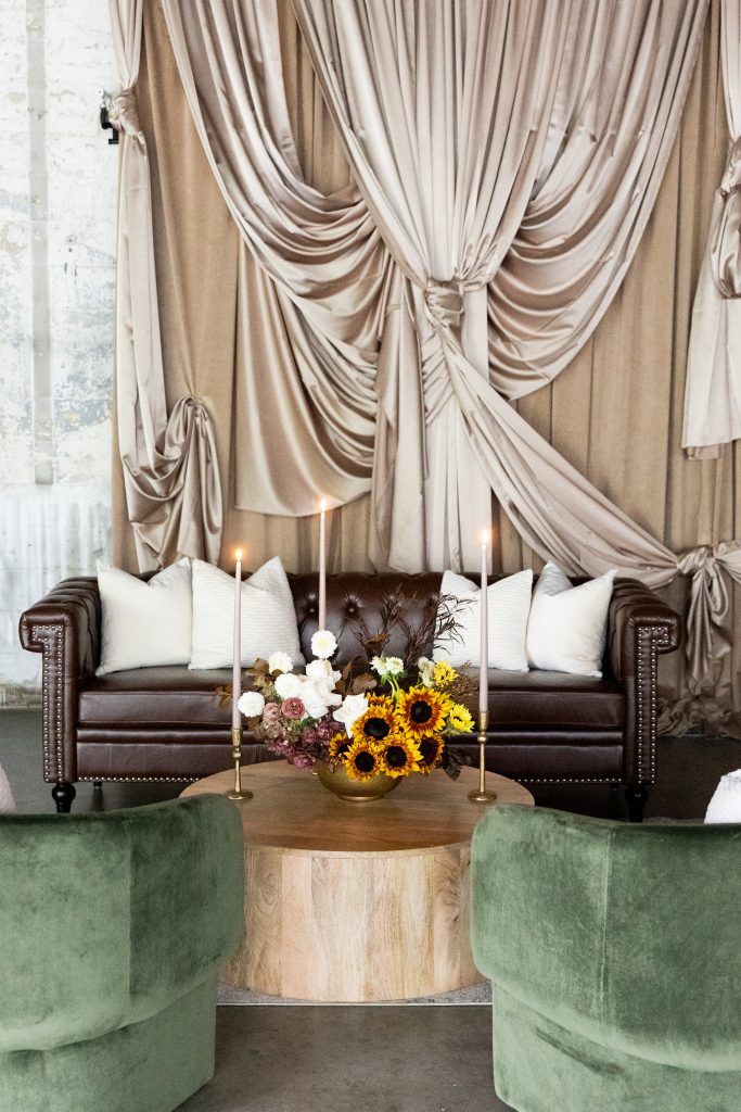 A lounge area with a leather sofa; velvet accent chairs; and a wooden, circular coffee table topped with a floral centerpiece and taper candles set against a designed drapery backdrop.