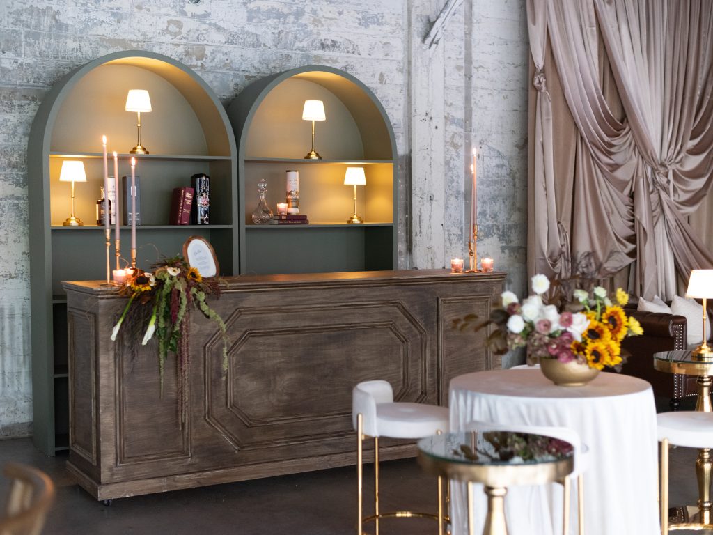 A wooden bar, with a spilling flower arrangement at one corner, set against curved shelving in the background and a high-top table with a floral centerpiece and seating in the foreground.