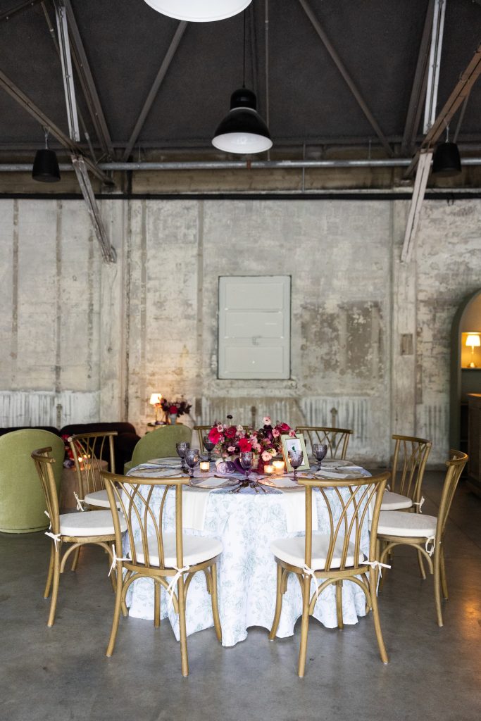 In a warehouse-style event space with high ceilings, distressed stone walls, and polished concrete floors, a round dining table setting with toile tablecloth, rustic-style chairs, and a floral centerpiece.