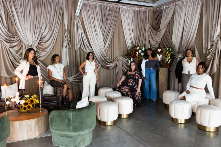 A group of women, posing individually and looking at the camera, dispersed throughout a staged setup of lounge areas against a designed drapery backdrop.