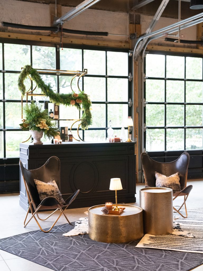 A wooden bar and gilded shelving decorated with a fir garland set against glass garage-style doors in the background and leather accent chairs, faux-fur rugs, and brass lounge tables in the foreground.