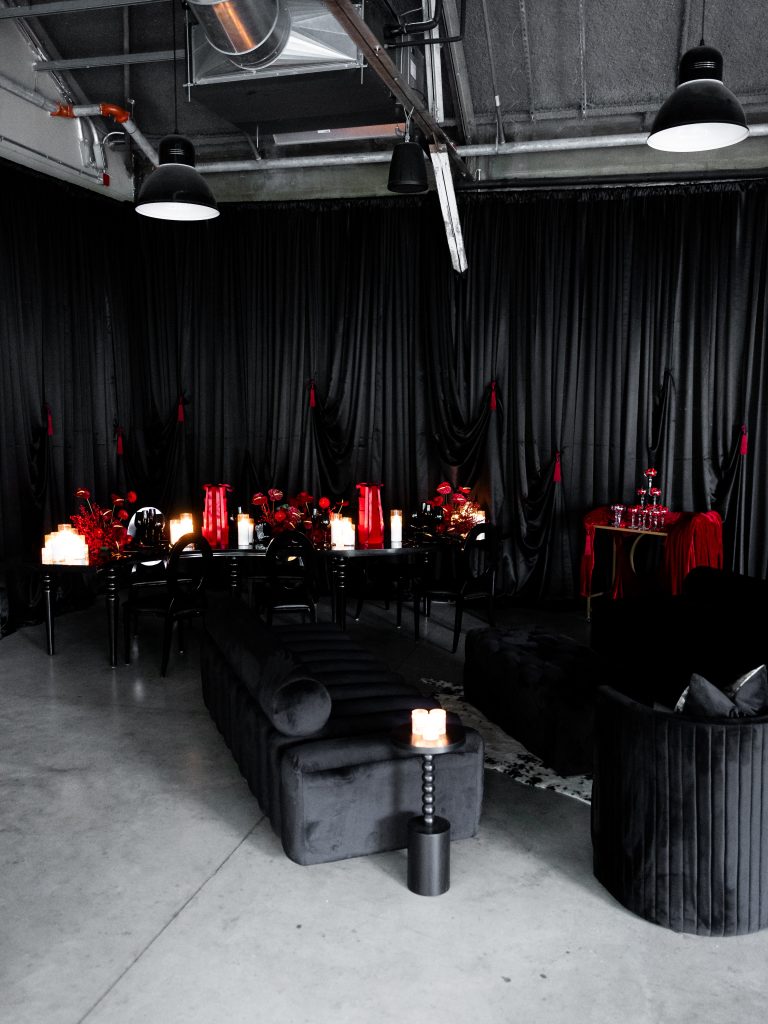 Black dining and lounge areas with pops of red floral arrangements and gift box decorations staged against a black designed drapery backdrop with red tassels in a modern-industrial venue featuring high ceilings and pendant lighting.