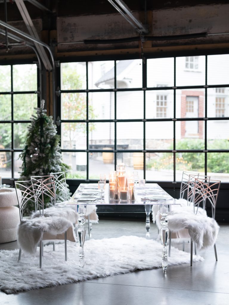A flocked Christmas tree in the background and an acrylic dining table lined with pillar candles and silver gallery chairs topped with faux sheepskin cushions in the foreground against large, glass garage-style doors.