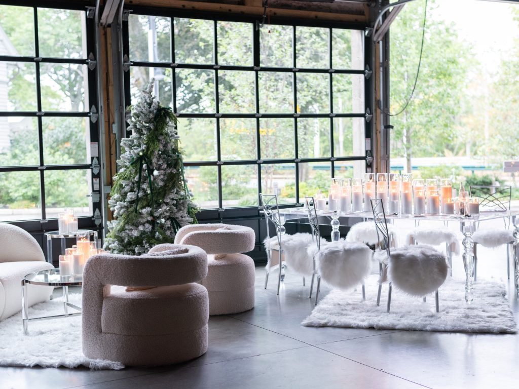 A flocked Christmas tree, a lounge area with all-white accent chairs, and an acrylic dining table filled with pillar candles and lined with silver gallery chairs topped with faux sheepskin cushions against large, glass garage-style doors.