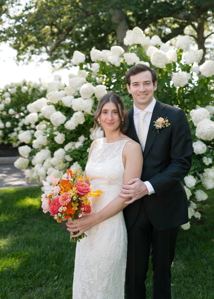 A bride, holding a bouquet, stands next to her groom in front of a large hydrangea bush.
