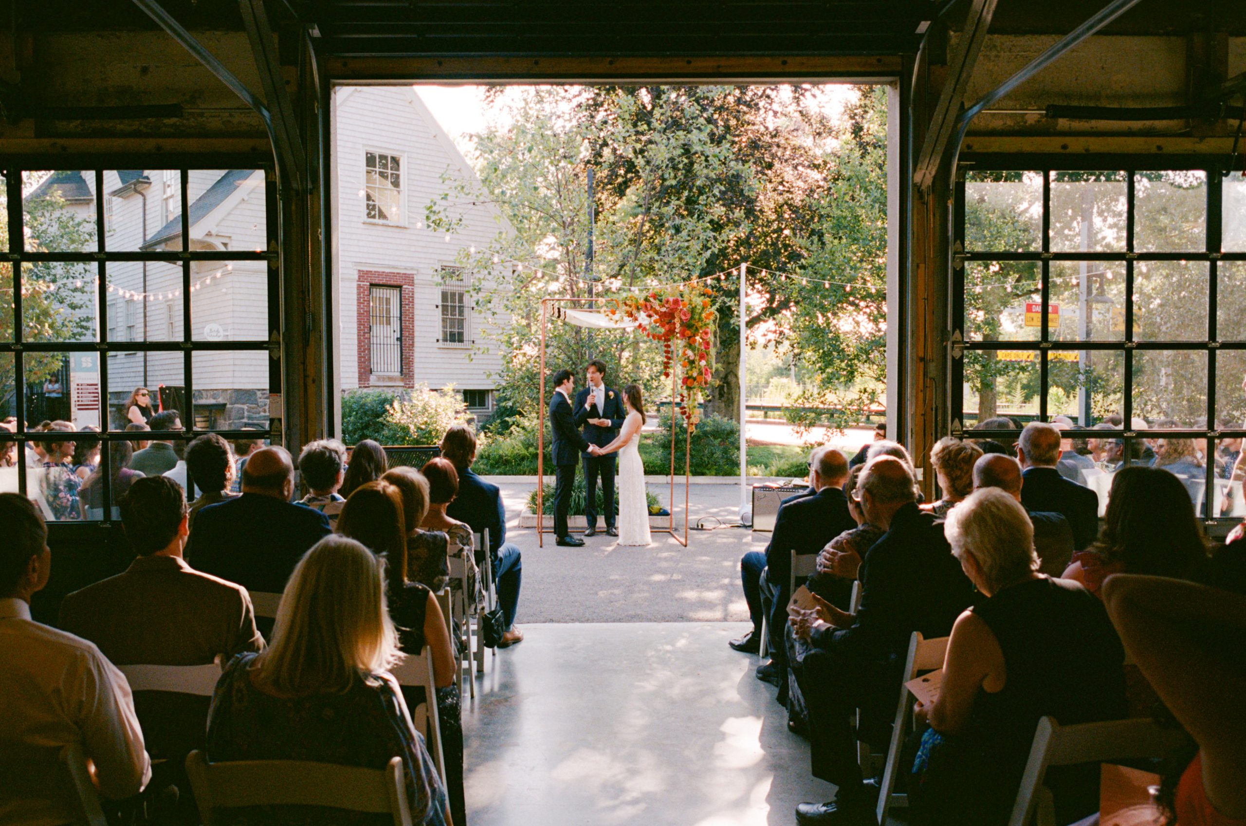 Wedding guests seated inside a venue looking upon an outdoor wedding ceremony -- including a bride, groom, and their officiant under a chuppah decorated with a spilling flower arrangement -- through a large, open overhead door.