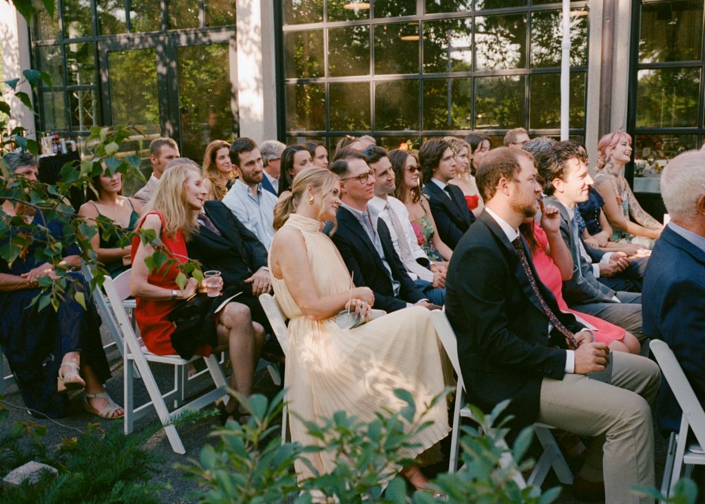 Rows of guests seated for an outdoor wedding ceremony in front of a series of large glass doors.