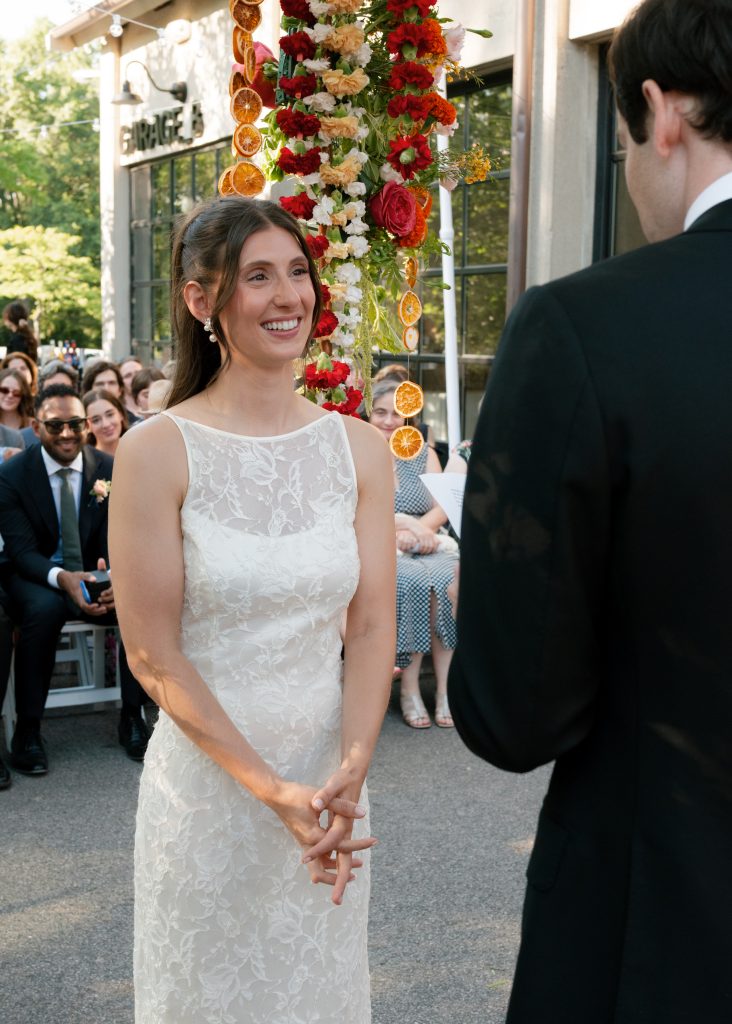 A smiling bride looks at her groom reading his vows under a spilling fresh and dried flower arrangement while their loved ones watch in the background in front of Garage B's building exterior and signage.