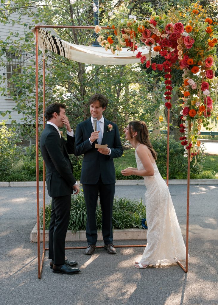 A groom and bride as well as their officiant laugh during an outdoor wedding ceremony under a chuppah decorated with a spilling arrangement of fresh and dried flowers.