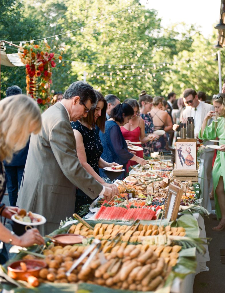 Wedding guests select hors d'oeuvres from a long grazing table during an outdoor cocktail hour.
