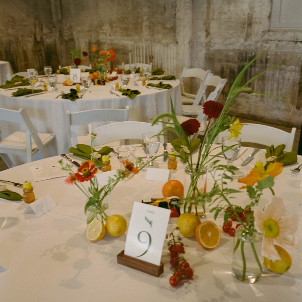 Two round banquet tables with white tablecloths and white folding chairs, decorated with sparse greenery and fresh fruits, against a distressed stone wall.