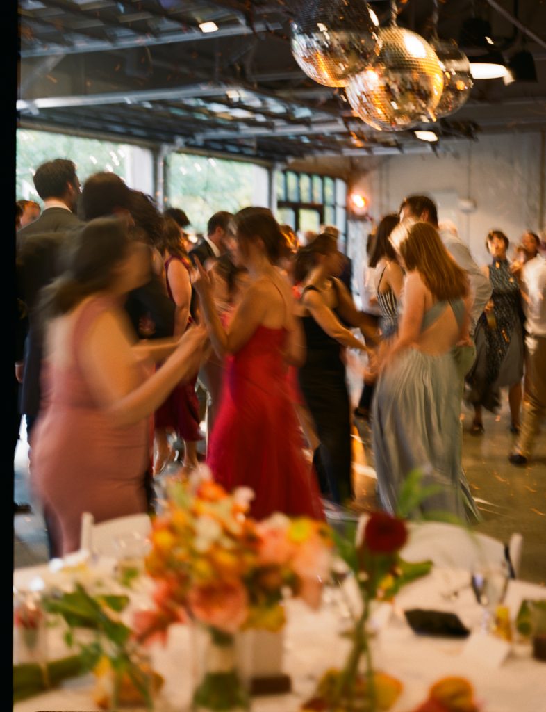 Wedding guests mingle and dance in a warehouse-style venue with disco balls installed at the ceiling and open overhead doors at one side.
