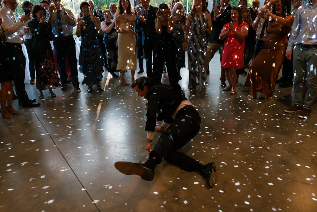 A groom breakdances while circled by loved ones watching with smiles underneath disco-ball lighting.