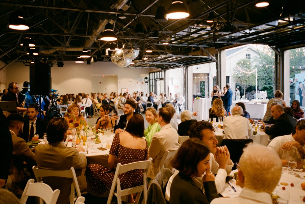Wedding guests seated at round banquet tables that fill a warehouse-style venue with disco balls installed at the ceiling and open overhead doors at one side.