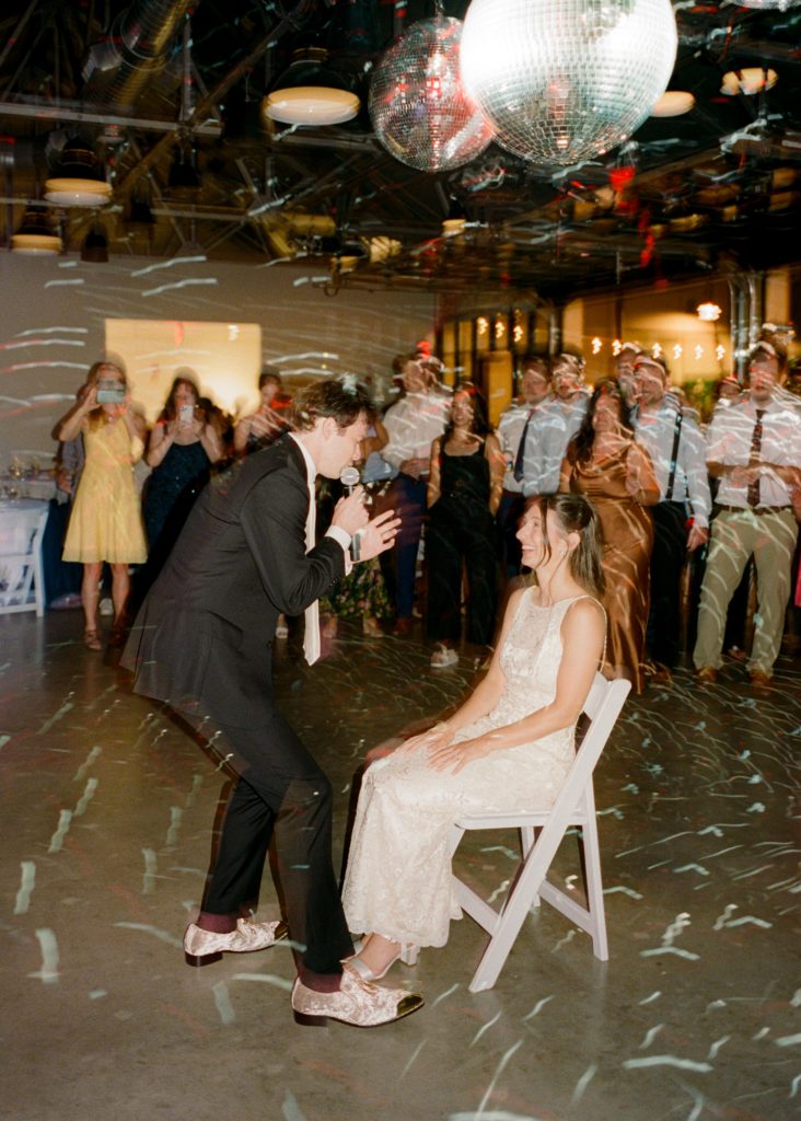 A groom serenades his smiling, seated bride while surrounded by loved ones underneath a series of disco balls.