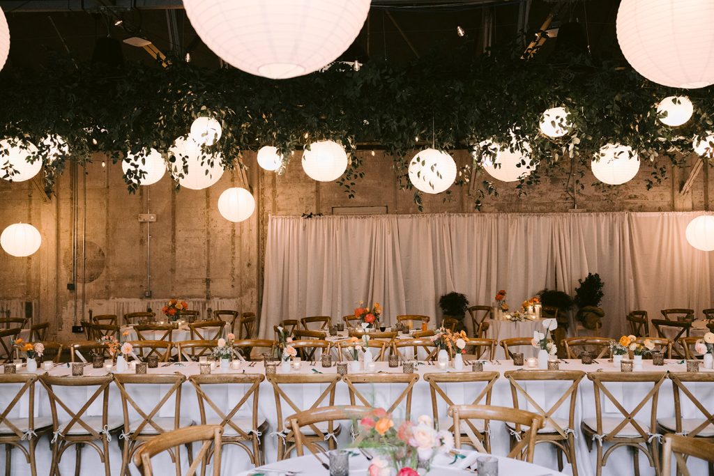 A ceiling arrangement of greenery and paper lanterns hangs over long banquet tables lined by rustic dining chairs at a wedding reception.