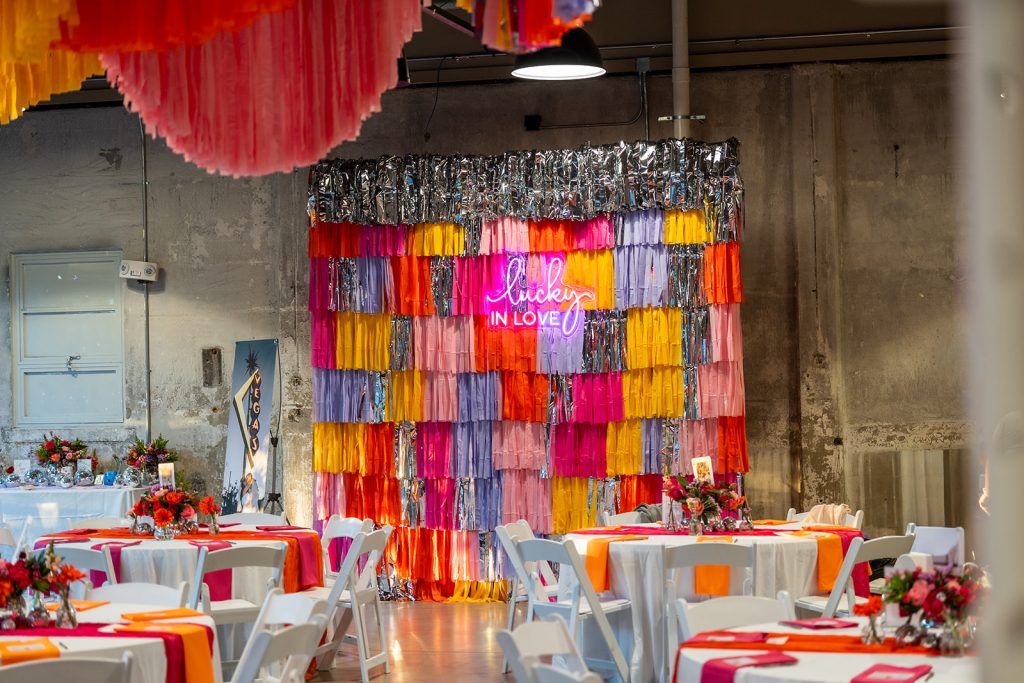 A series of round banquet tables in the foreground with a photo backdrop comprised of layers of brightly-colored streamers and a neon sign that reads "Lucky in Love" in the background.