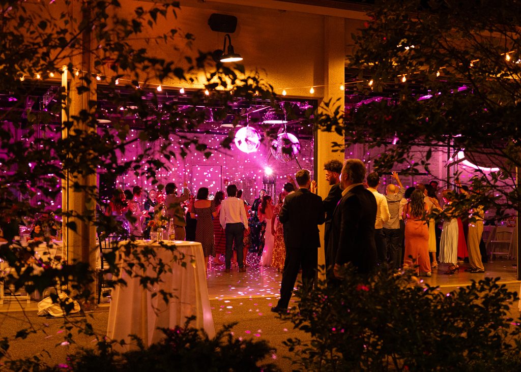 A view from a patio into a wedding celebration beyond open floor-to-ceiling doors at a modern-industrial venue.