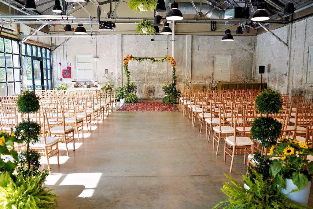A warehouse-style wedding venue -- with distressed stone walls, polished concrete flooring, large glass windows, and pendant lighting -- set up for a wedding ceremony that includes rows of wooden Chivari chairs, an arch that features clusters of greenery and sunflowers, a vintage rug, and potted greenery and shrubs as décor.