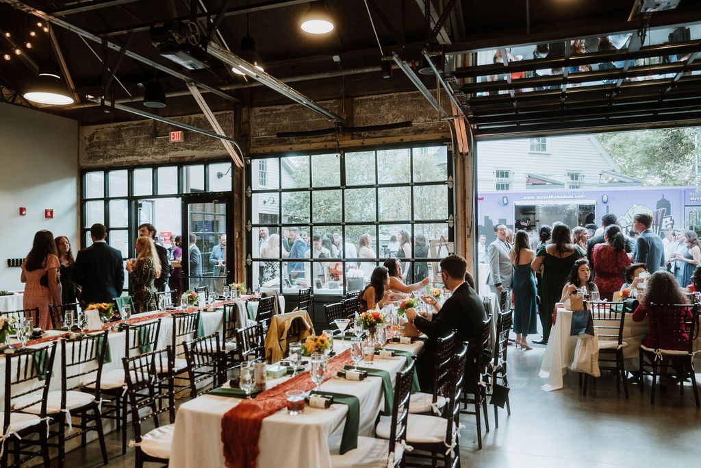 A crowd of wedding guests mingles both inside a venue at rectangular banquet tables as well as on an outdoor patio beside a food truck, separated by a bank of large, glass overhead garage-style doors.