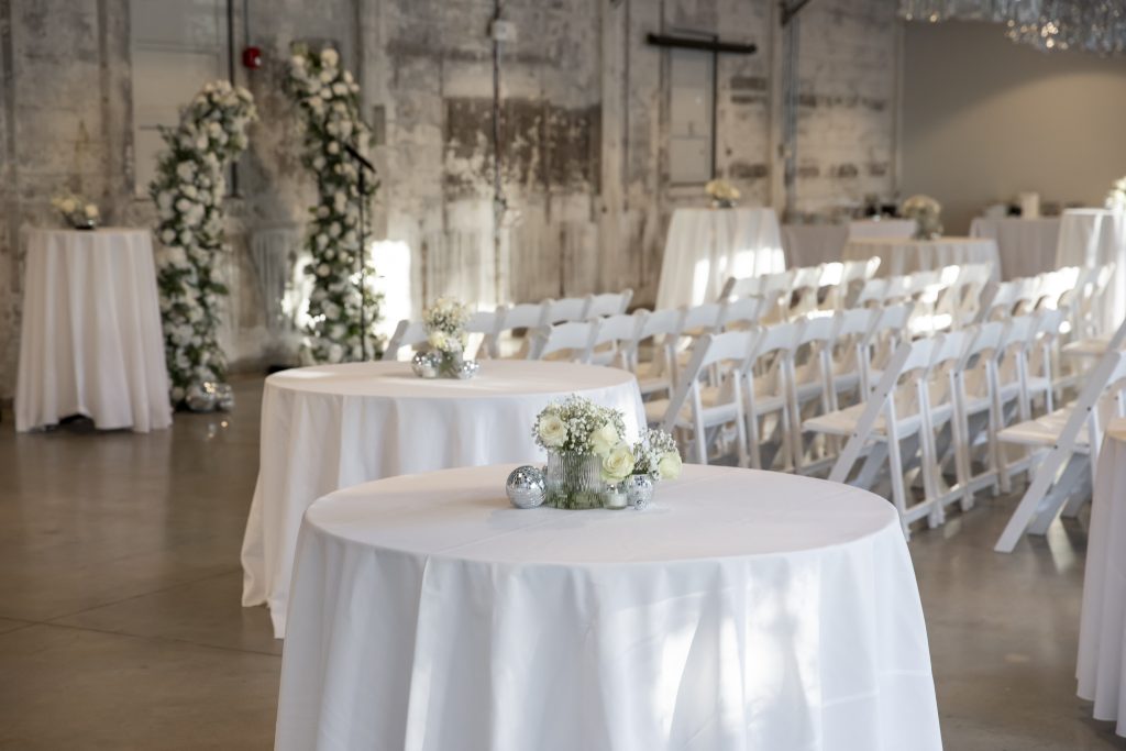 An all-white wedding ceremony setup set against distressed stone walls that includes a floating flower arch, rows of folding chairs, and high-top tables.