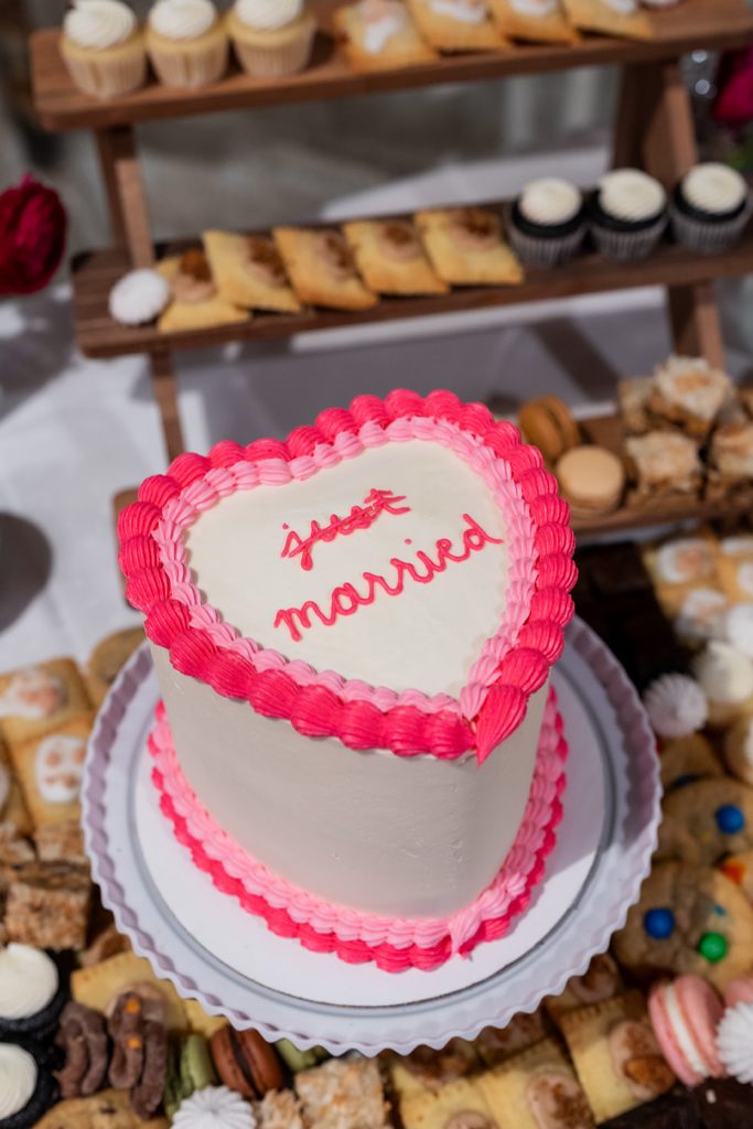 A small heart-shaped cake, sitting atop a display of assorted cookies, that reads "Just Married" with the word "just" stylized with a strikethrough.