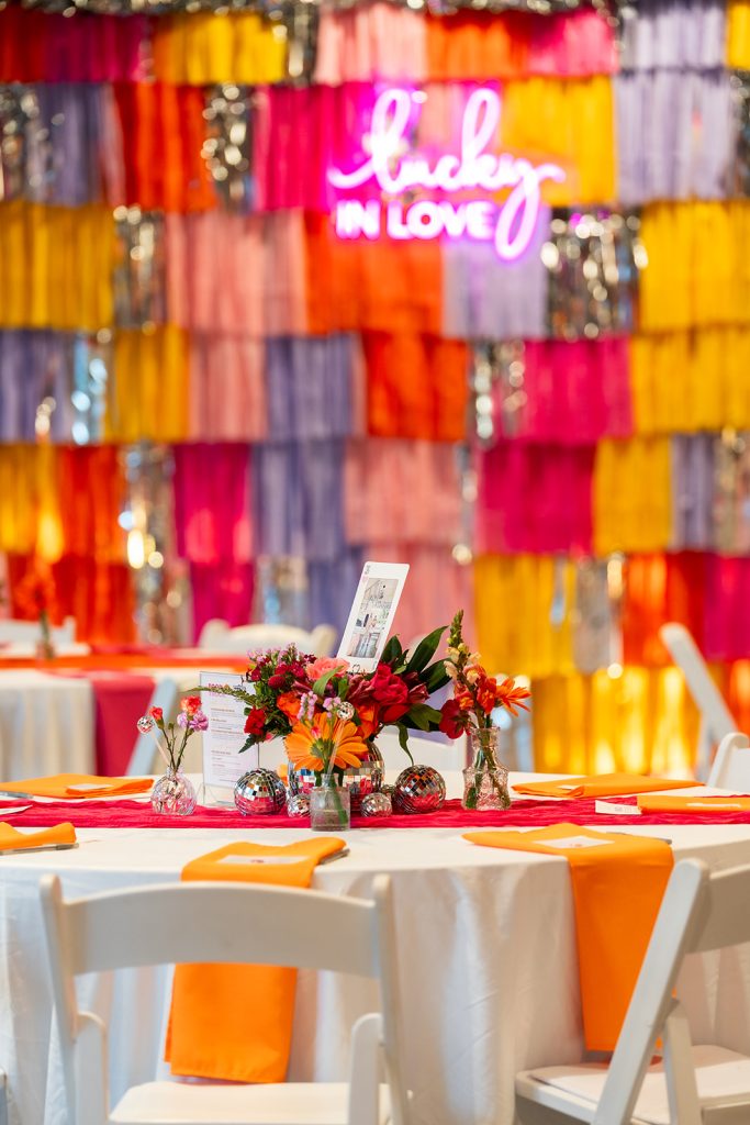 A round banquet table with a centerpiece consisting of bright florals and disco balls set against a backdrop made of colorful streamers and a neon sign that reads "Lucky in Love".