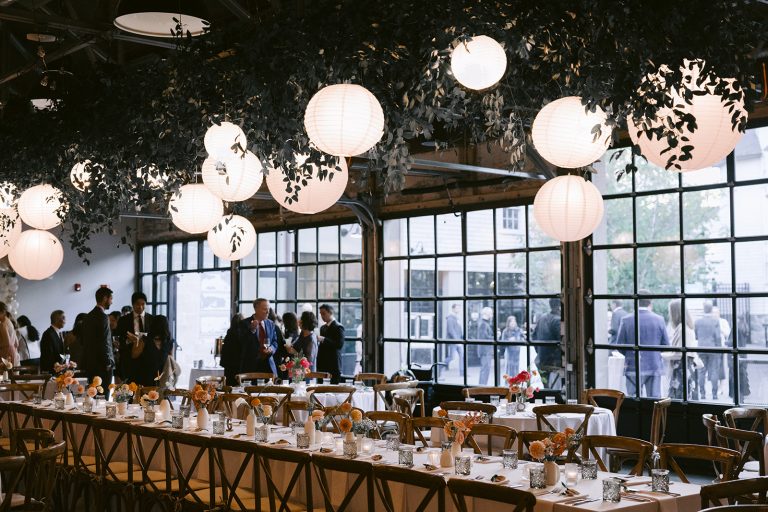 Several people mingle around a long rectangular table and a series of round banquet tables decorated with floral bud vases under a ceiling arrangement of greenery and paper lanterns inside a wedding venue while other people mingle outside on an adjacent patio, seen through a set of floor-to-ceiling glass overhead doors.