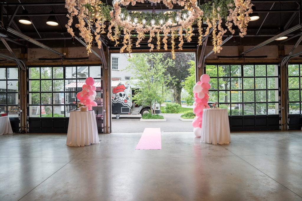 A view through a bank of large, garage-style glass doors from inside an event venue that features a ceiling arrangement of hanging cherry blossoms and greenery as well as two high-top tables and two pink balloon column structures to an adjacent outdoor patio at which a food truck is parked.