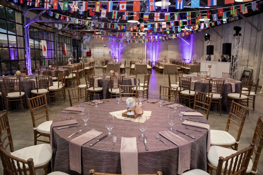 A warehouse-style venue outfitted with streamers of flags at the ceiling, banquet tables featuring globe centerpieces, and a DJ booth that features a bank of large glass windows through which a tented area can be seen.
