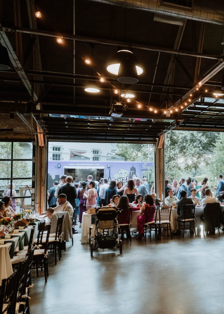 Wedding guests mingle at banquet tables indoors and at a food truck outdoors through an open overhead glass door.