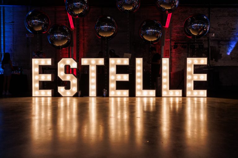 The name "Estelle" stylized as a large, lighted monogram backed by metallic balloons and red uplighting.