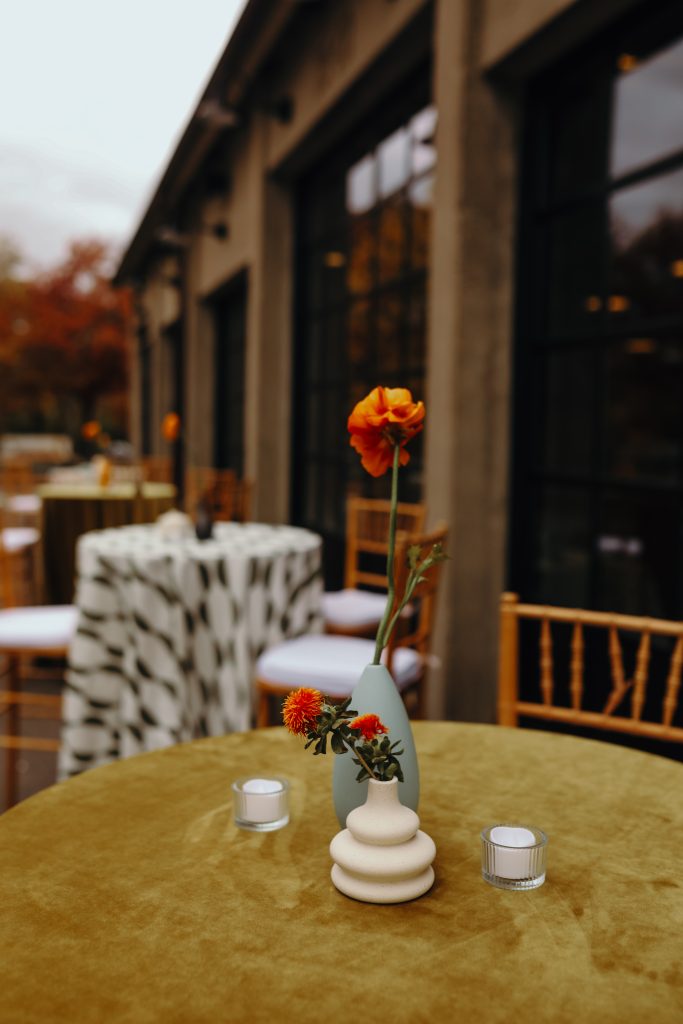 Minimalist floral bud vases sit atop a velvet tablecloth in the foreground with a series of other high-top tables draped in patterned linens against a modern-industrial building exterior in the background.