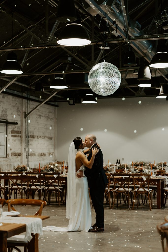 A groom and bride dance privately under a disco ball in a warehouse-style wedding venue outfitted with rustic tables and chairs.