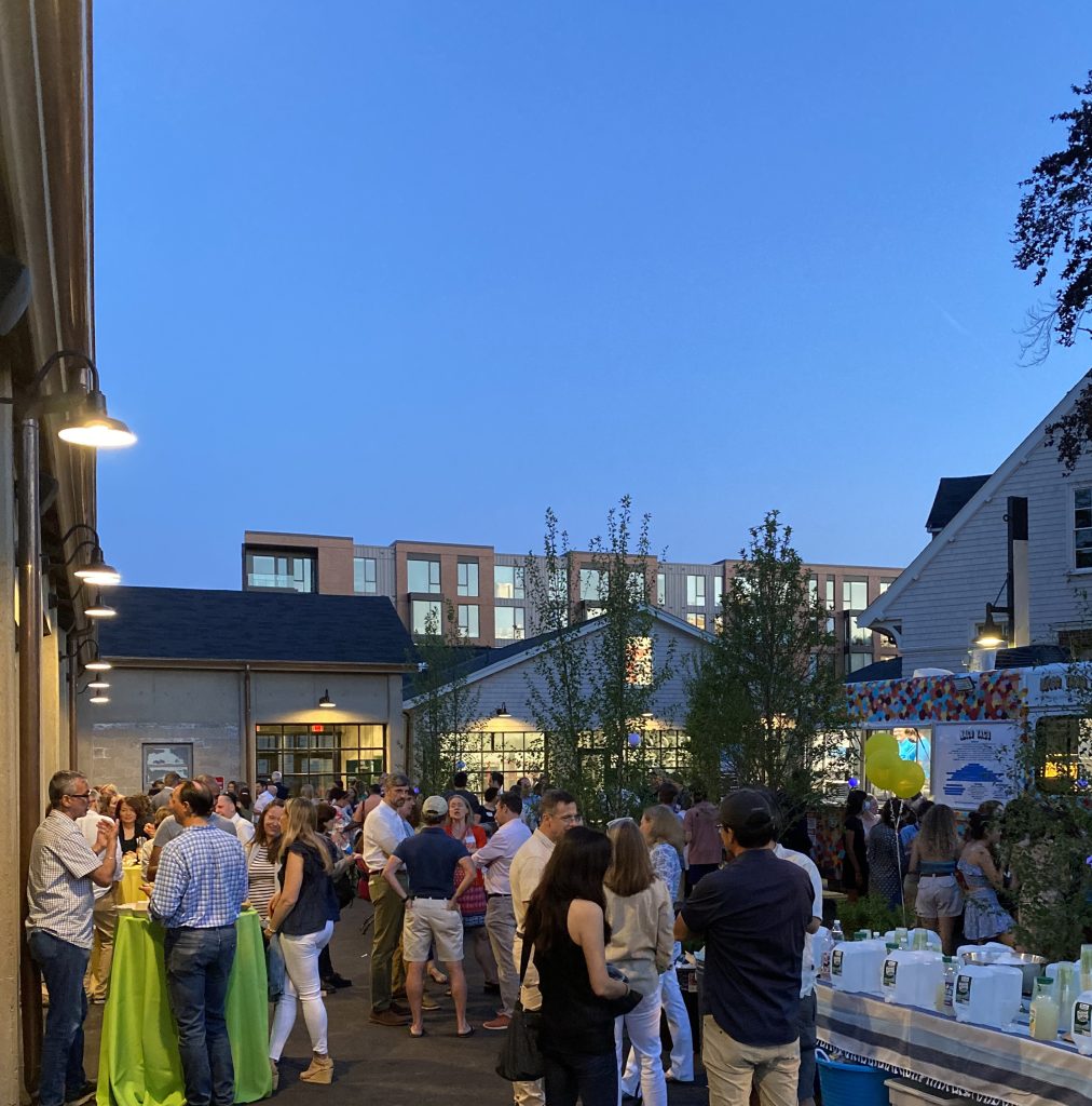 A crowd of casually-dressed parents gathers on an outdoor patio around a series of high-top tables while their children order tacos from a nearby food truck.