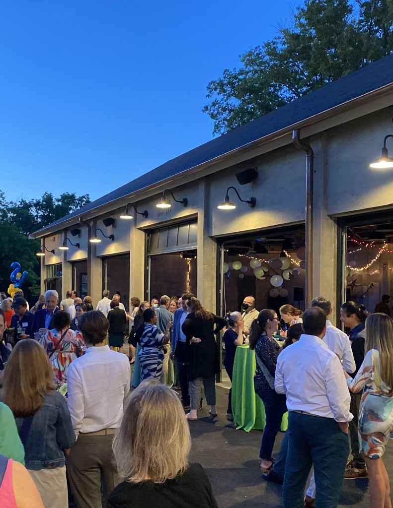 A crowd of casually-dressed parents gathers on an outdoor patio around a series of high-top tables adjacent to a building featuring a series of open garage-style doors.
