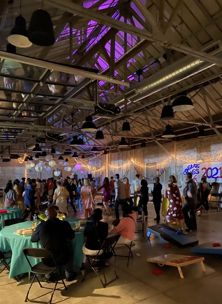 Guests at a graduation party for the Class of 2021 fill a modern-industrial event venue among round banquet tables and lawn games.