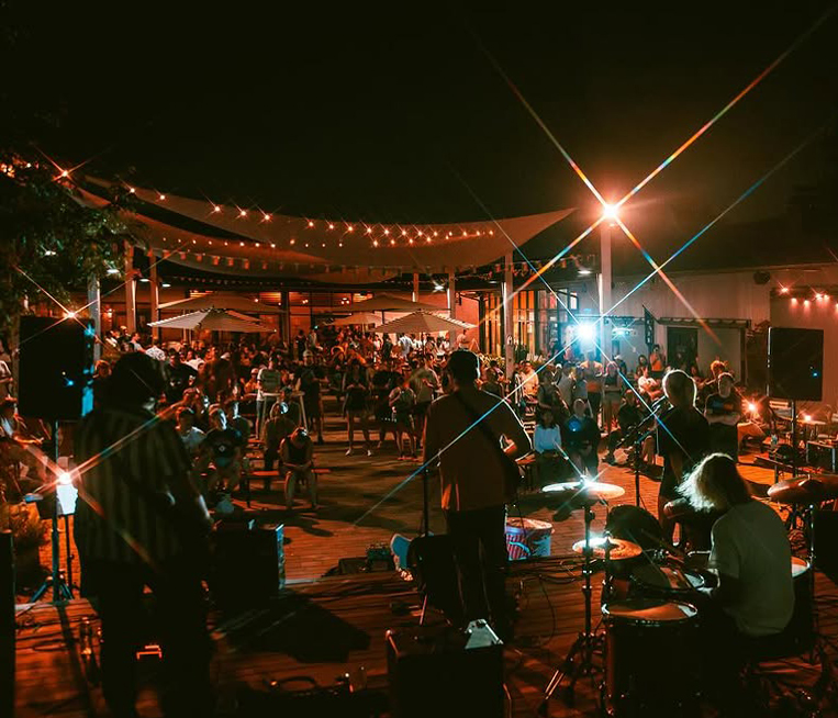 A view from behind a band performing on an outdoor stage to a crowd gathered in a courtyard.